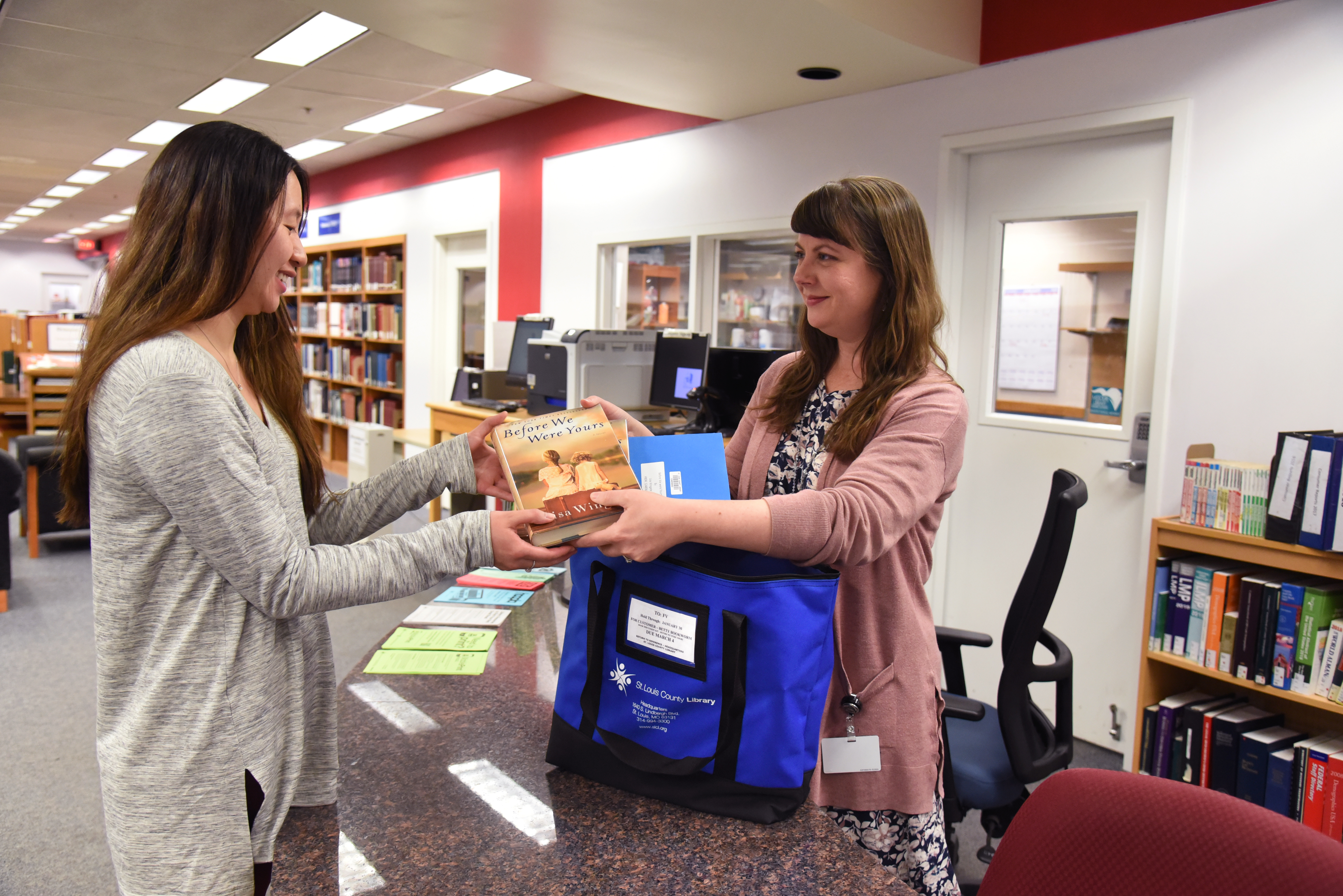 A smiling female librarian behind a desk handing a female patron a couple books out of a blue book discussion kit bag.