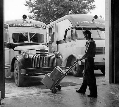 A white male library employee wheeling two boxes out of a garage opening towards one of two nearby parked 1940s bookmobile trucks