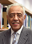 Lynn Beckwith, Jr., Ed.D. standing by a shelf of library books smiling and wearing a gray stripped suit and silver tie