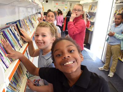 A group of kids smiling and flipping through books on the shelves inside a bookmobile