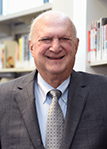 Ted Sanditz in front of library bookshelves smiling wearing a gray suit coat light blue shirt and gray tie