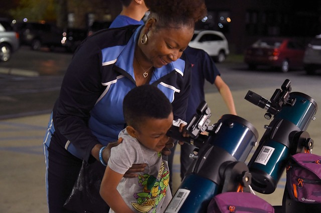 A black mother holds young boy up to a telescope setup for viewing on parking lot