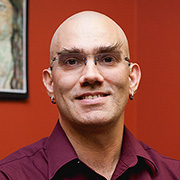 Crom Saunders smiling wearing glasses and a maroon button down dress shirt against red painted walls.