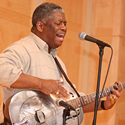Rev. Robert B. Jones, Sr. performing on stage at a microphone while playing a silver guitar