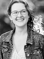 Emma Mills black and white headshot of her outside, the breeze and light coming through her long curled hair and she is wearing glasses with a jean jacket