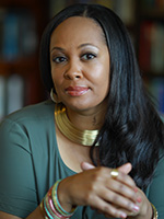 Kim Johnson headshot with her looking at the camera wearing a large gold necklace and a teal long sleeve top with her hands folded in front of her