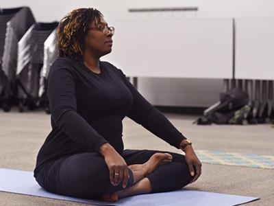 A black woman with glasses dressed in all black exercise clothing sits in a yoga position on a light colored mat