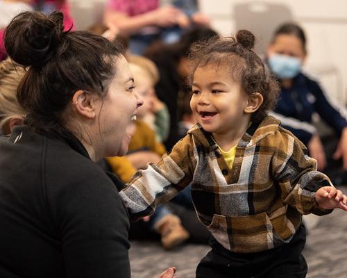 A smiling toddler standing in front of his mother who is smiling with the mouth open at a library program