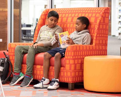 Two boys sitting on a bright orange couch talking and looking at a book